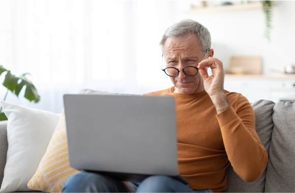 An elderly man squinting while looking at his laptop, adjusting his glasses to see more clearly.